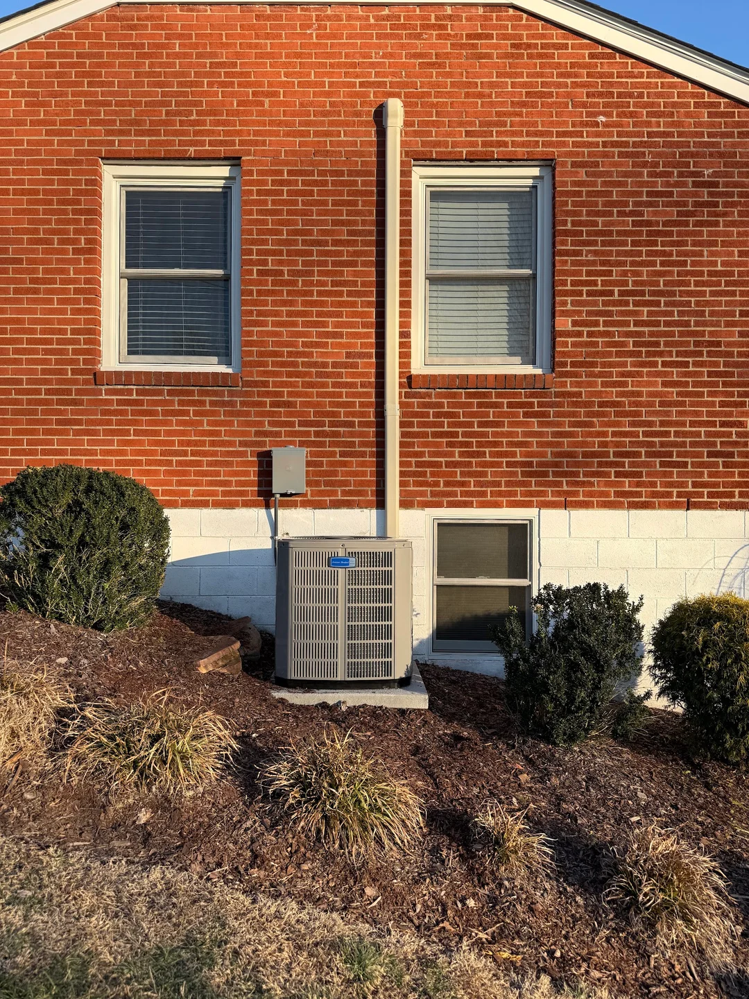 A split-system condenser on a pad beside a red-brick house with a painted block foundation